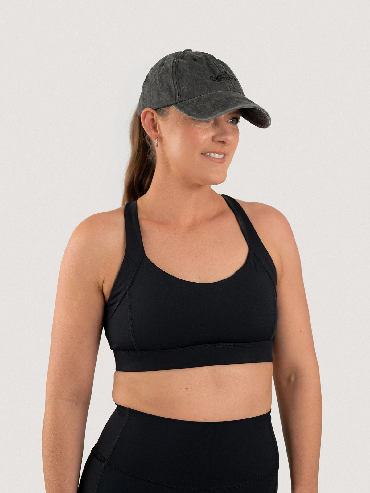 Woman wearing the Aella Eye Off-Duty Hat in Ash with a black Aella Storm Hybrid Sports Bra.